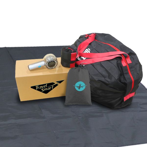Black foldable duffel bag with red trim beside a kraft box, a travel adapter and a luggage tag on a black cloth