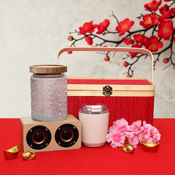 Red patterned box with a wooden lid beside a glass jar, a white tumbler and a wooden speaker on red cloth with blossoms