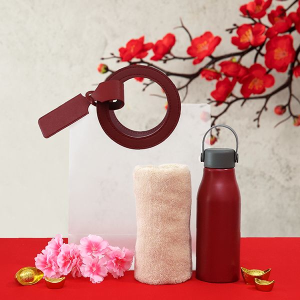 White tote bag, maroon bag charm, rolled towel and maroon bottle arranged on red cloth under cherry blossom branches