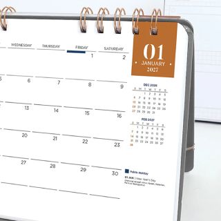 Monthly calendar page standing on a desk, with a dated grid, a coloured date tab and small reference months.