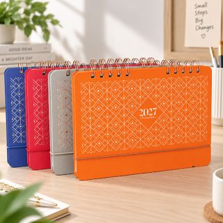 Four wire-o desk calendars lined up on a wooden desk in blue, red, grey and orange beside a plant.