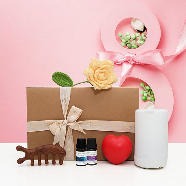Kraft gift box tied with ribbon beside a wooden comb, essential oil bottles, a red stress ball and a white diffuser on a pink backdrop